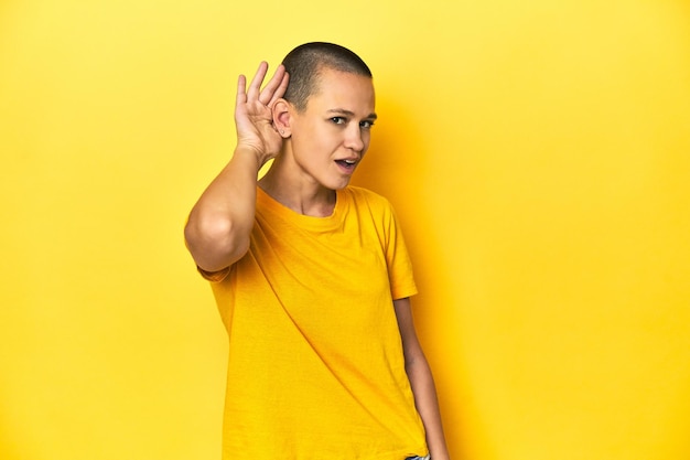 Junge Frau in gelbem T-Shirt, gelbem Studiohintergrund, der versucht, einem Klatsch zuzuhören