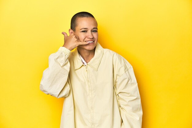 Junge Frau in gelber Jacke, Studio-Hintergrund zeigt eine Handy-Anrufgestur mit Fingern