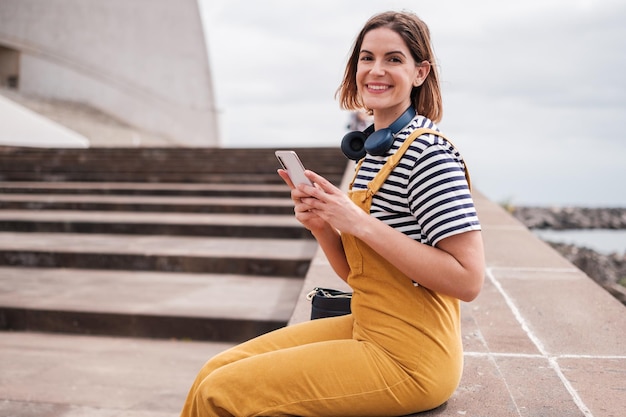 Junge Frau in gelber Latzhose mit dem Smartphone und lächelnden Blicken auf die Kamera Konzept Lifestyle im Freien soziale Netzwerke