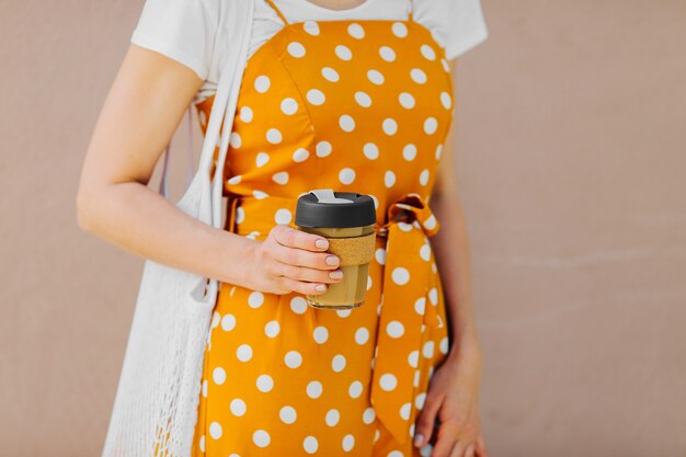 Junge Frau in gelber Sommerkleidung hält wiederverwendbare Kaffeetasse. Nachhaltiger Lebensstil.