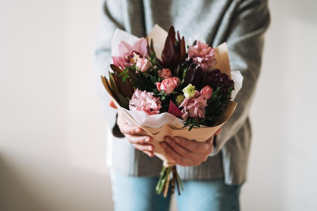 Junge Frau in grauem Strickpullover mit Blumenstrauß aus verschiedenen rosafarbenen Blumen in den Händen auf grauem Hintergrund, selektiver Fokus