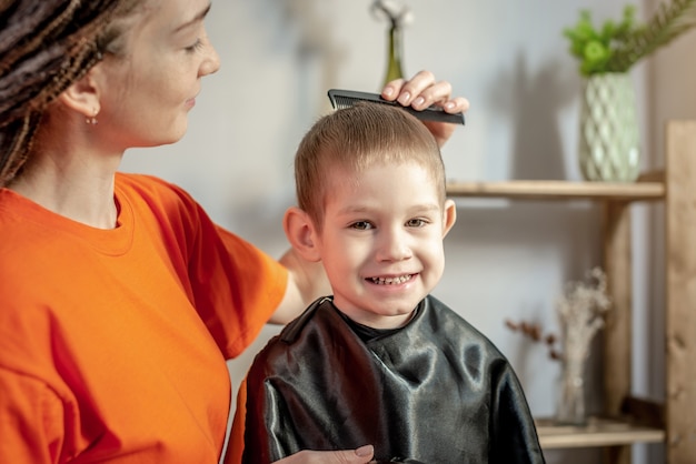 Junge Frau in hellen Kleidern kämmt die Haare eines kleinen Jungen, bevor sie sie schneidet