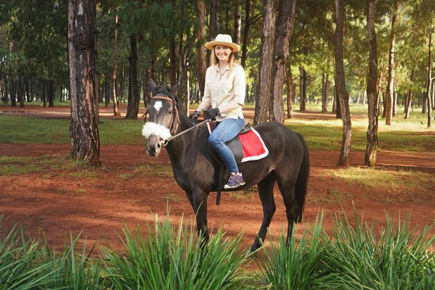 Junge Frau in Hemd und Strohhut, die auf einem dunkelbraunen Pferd im Park reitet, verschwommen hohe Kiefern im Hintergrund