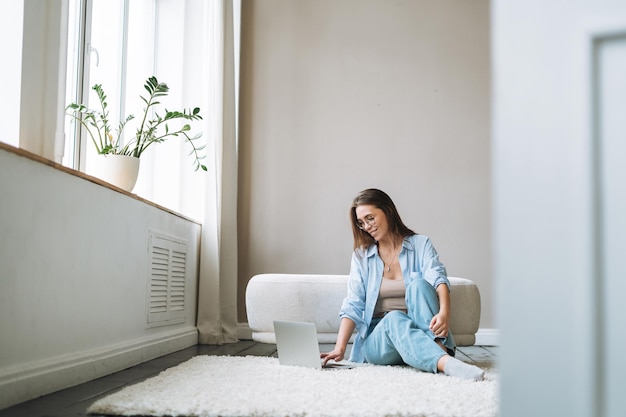 Junge Frau in Jeans, die am Laptop arbeitet und zu Hause auf dem Teppich sitzt, Blick von den Türen