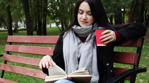 Junge Frau in Jeans, Mantel und Schal, auf einer Parkbank. Eine Frau liest allein im Freien ein Buch und trinkt Kaffee oder ein anderes heißes Getränk.