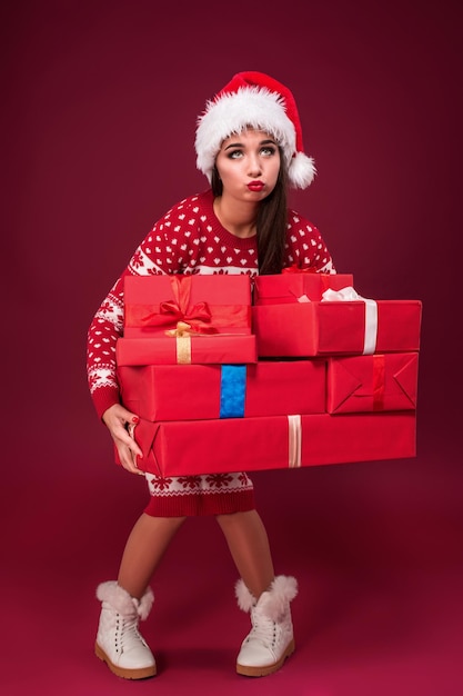 Junge Frau in Kleid und Weihnachtsmütze mit Geschenkboxen auf rotem Hintergrund