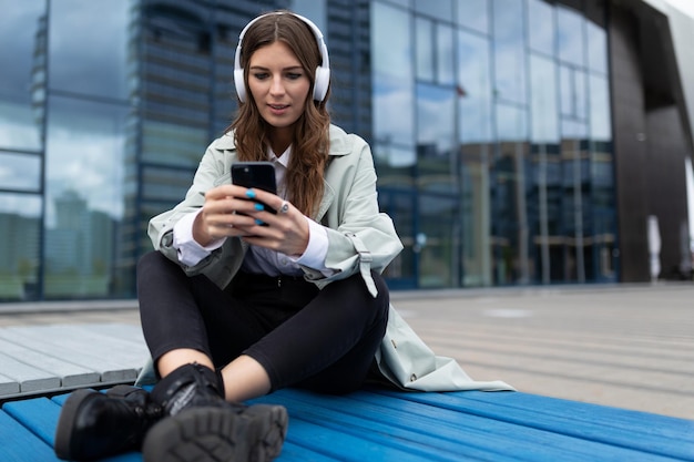 Junge frau in kopfhörern mit einem handy, das musik mit gekreuzten beinen hört, sitzt neben dem