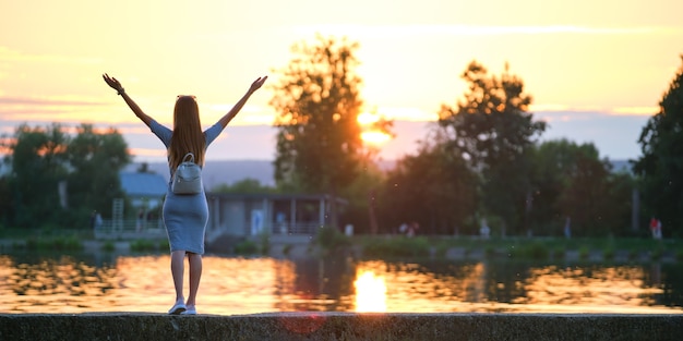 Junge Frau in lässigem Outfit, die sich am See mit erhobenen Händen an einem warmen Abend entspannt. Erfolgs- und Wellnesskonzept.