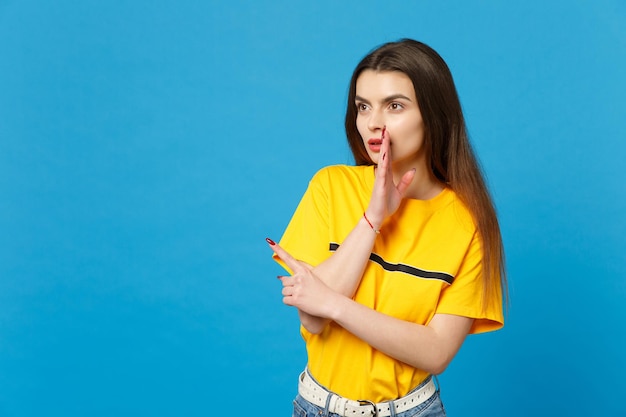 Junge Frau in lebendiger Freizeitkleidung flüstert Klatsch und erzählt mit Handgesten Geheimnisse, zeigt mit dem Zeigefinger isoliert auf blauem Hintergrund im Studio. People-Lifestyle-Konzept. Mock-up-Kopienbereich.