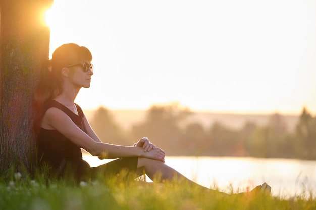 Junge Frau in legerem Outfit sitzt auf grünem Rasen und entspannt sich an warmen Abenden am Seeufer Sommerferien und Reisekonzept