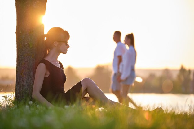 Junge Frau in legerem Outfit sitzt auf grünem Rasen und entspannt sich an warmen Abenden am Seeufer Sommerferien und Reisekonzept