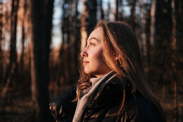 Junge Frau in Mantel und Schal, die im Herbstwald steht und den Sonnenuntergang im Freien betrachtet