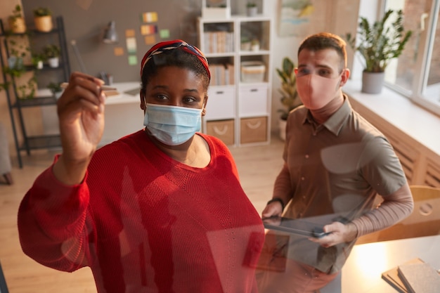 junge Frau in Maske, die auf die Glaswand schreibt, die sie zusammen mit Mann im Büro über Projekt arbeiten