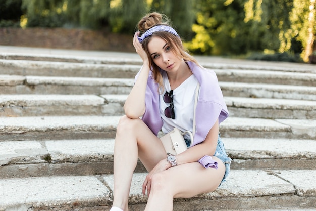 Junge Frau in modischer Denim-Freizeitkleidung mit lila Bandana und Sweatshirt sitzt auf den Stufen