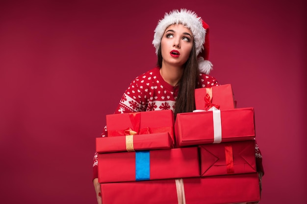 Junge Frau in Pullover und Weihnachtsmütze mit Geschenkboxen auf rotem Hintergrund