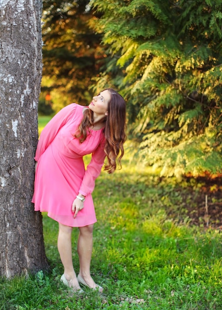 Junge Frau in rosafarbenem Kleid, an Baum gelehnt, nach oben schauend, mit Sonnenuntergangslichtpark im Hintergrund.