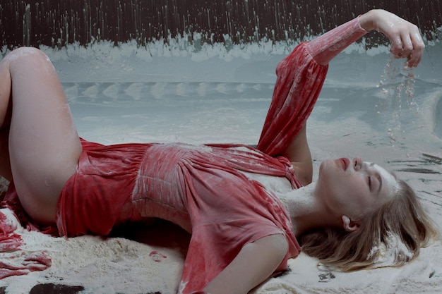 Junge Frau in rotem Kleid liegt in Mehl auf dem Boden und gießt Mehl auf sich