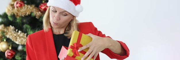 Junge Frau in rotem Weihnachtsmann-Hut öffnet Neujahrsgeschenk in der Nähe von Baum und ist überrascht von der Auswahl