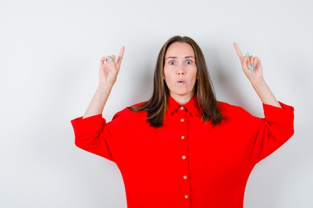 Foto junge frau in roter bluse, die mit den fingern nach oben zeigt und schockiert aussieht, vorderansicht.