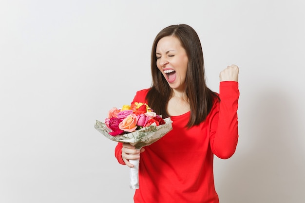 Junge Frau in roter Kleidung, die einen Strauß schöner Rosenblumen hält und die Siegergeste tut, die auf weißem Hintergrund lokalisiert wird. Valentinstag oder Internationaler Frauentag, Geburtstag, Urlaubskonzept.