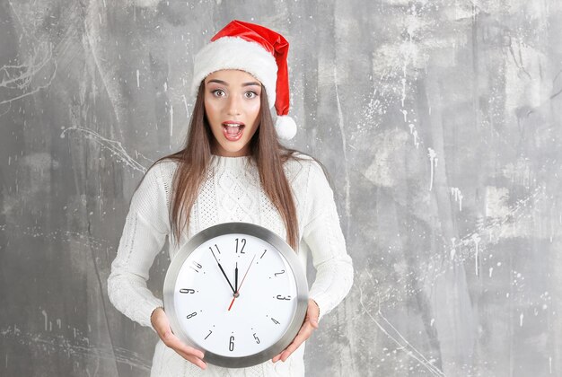 Junge Frau in Santa Hut mit Uhr auf Grunge-Hintergrund. Weihnachts-Countdown-Konzept