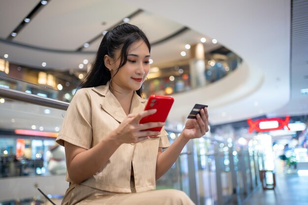 Foto junge frau in schlauer freizeitkleidung, die viele papier-shopping-taschen trägt und mit kreditkarte im einkaufszentrum online mit dem handy einkaufen geht