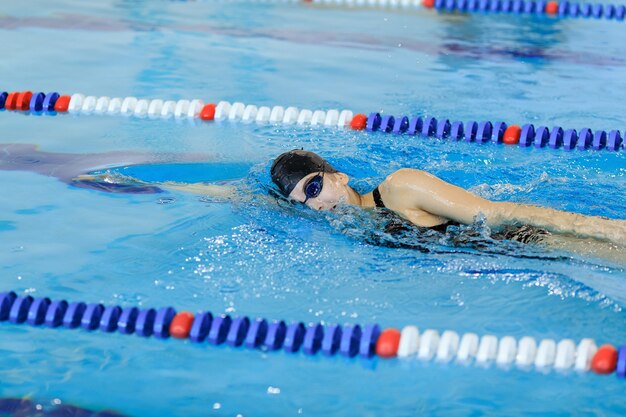 Junge Frau in Schutzbrillen und Mütze schwimmen im vorderen Crawl-Stroke-Stil im blauen Wasser-Indoor-Rennpool.