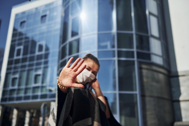 Junge Frau in Schutzmaske, die bei sonnigem Tag im Freien in der Nähe des Geschäftsgebäudes steht Konzeption des Coronavirus