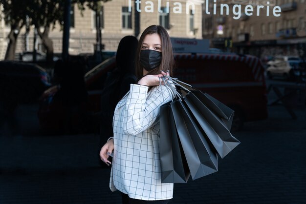 Junge Frau in Schutzmaske hält viele Einkaufstüten auf schwarzem Hintergrund. Konzept des schwarzen Freitags.
