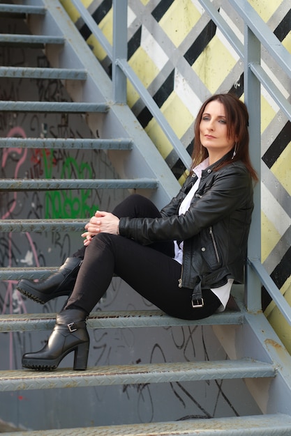 Junge Frau in schwarzem engem Denim, weißem Hemd und Lederjacke sitzt auf Metalltreppe