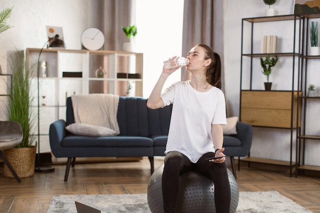 Junge Frau in schwarzen Leggings und T-Shirt mit Smartphone sitzt auf fit Ball Trinkwasser