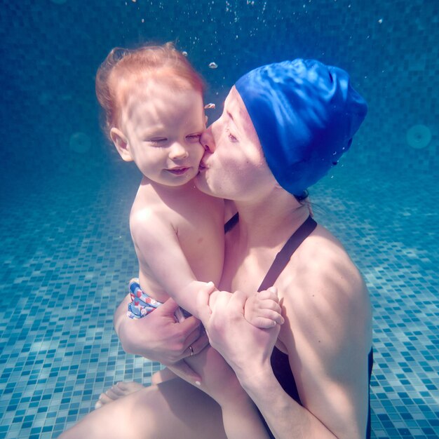 Foto junge frau in schwimmmütze hält ein süßes kind in den armen und küsst das baby auf die wange charmante dame sitzt auf dem schwimmbadboden unter wasser und küsst ihren sohn konzept des schwimmens familie und mutterliebe