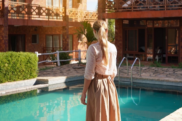 Junge Frau in Sommerkleid und Rock blickt in den leeren Hotelresortpool, Blick von hinten