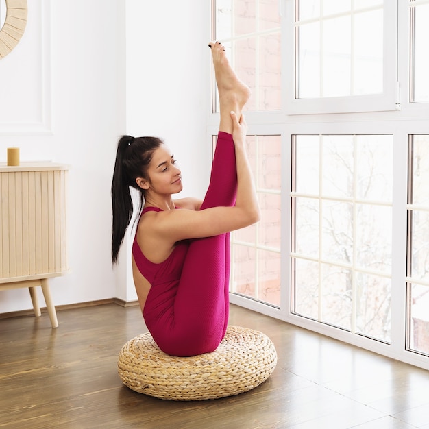 Junge Frau in Sportbekleidung, die Yoga praktiziert, sitzt auf dem Boden und macht Urdhva Mukha Paschimottanasana Übung in einem Raum nahe einem großen Fenster