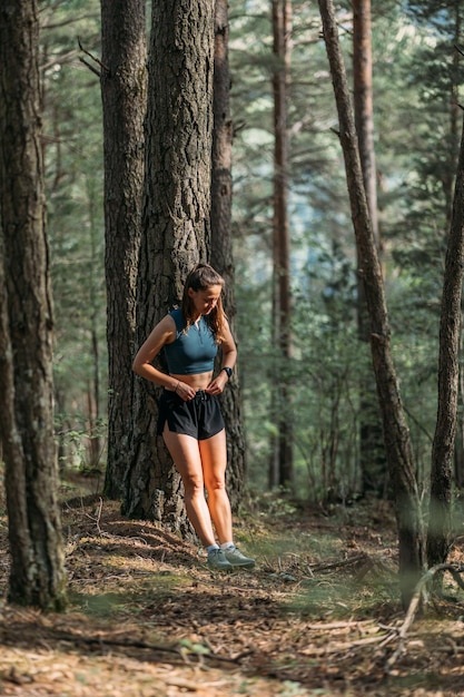Junge Frau in Sportbekleidung lehnte sich an einen Baum und glättet die Kleidung im Wald während des Trainings Trailrunning und aktives Lebenskonzept