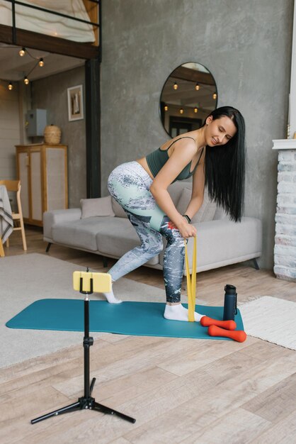 Junge Frau in Sportbekleidung macht eine Übung mit einem elastischen Band und nutzt ein Smartphone auf einem Stativ zu Hause im Wohnzimmer für Videounterricht Das Konzept von Sport und Erholung