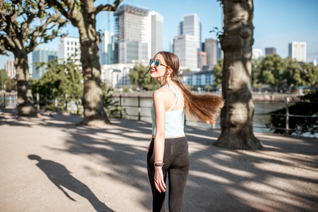 Junge Frau in Sportkleidung, die im Park mit Wolkenkratzern im Hintergrund in der Frankfurter Stadt spaziert und zurückblickt?