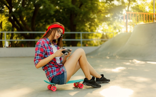 Junge Frau in stilvoller Kleidung sitzt auf einem Skateboard und verwendet an einem sonnigen Tag eine Retro-Kamera im Skatepark. Jugendkonzept