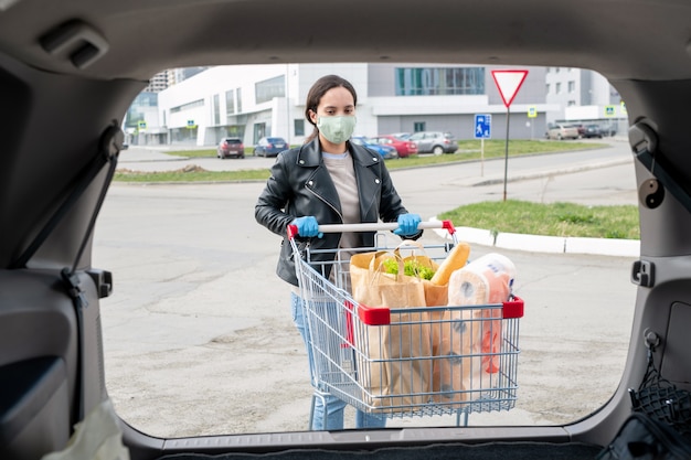 Junge Frau in Stoffmaske und Handschuhen, die Einkaufswagen zum Auto schieben, um Produkte in Kofferraum zu laden