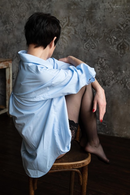 Junge Frau in Strümpfen und blauem Oversize-Hemd sitzt auf Stuhlbeinen hochgezogen und mit Blick auf die Wand