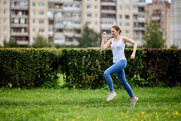 Junge Frau in Tanktop und Leggings joggt im öffentlichen Park neben einem Wohngebiet mit Wohnhochhäusern