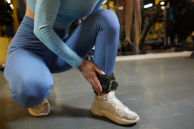 Junge Frau in Turnschuhen, die sich einen Lederriemen an den Knöchel schnallt, während sie auf dem Boden eines Fitnessstudios in einem sitzt