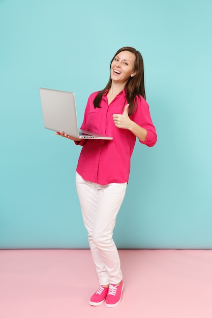 Junge Frau in voller Länge in rosa Hemdbluse, weiße Hose mit Laptop-PC isoliert auf hellrosa blauer Pastellwand..