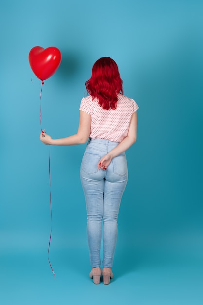 junge Frau in voller Länge steht mit dem Rücken und hält einen roten fliegenden Ballon in Form eines Herzens