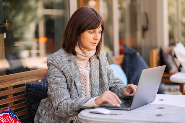 Junge Frau in warmem Pullover und Blazer, die im Café auf der Terrasse am Netbook arbeitet. Herbstmorgen. Streetstyle.