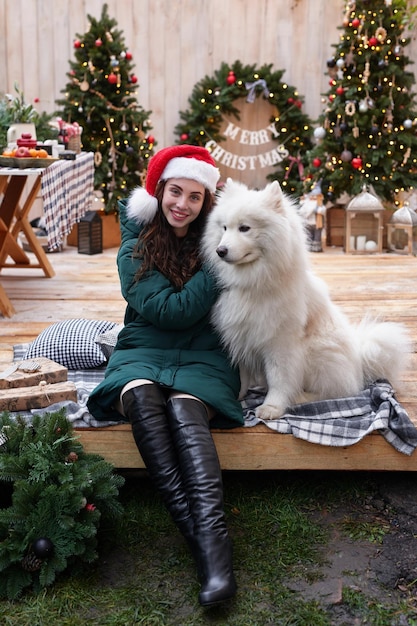 Junge Frau in Weihnachtsmütze auf dem Hintergrund des Weihnachtsbaums mit weißem Samoyed-Hund im Freien Hofdekoration für das neue Jahr