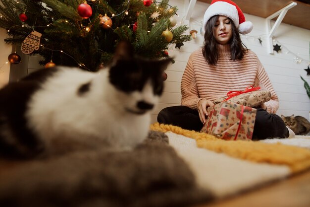 Junge Frau in Weihnachtsmütze öffnet Weihnachtsgeschenk mit ihrer Katze unter Weihnachtsbaum im festlichen Raum