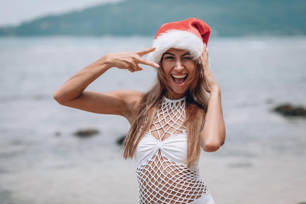 Junge Frau in weißem Badeanzug und Weihnachtsmütze am tropischen Strand lächelt in die Kamera. Weihnachtsferien. Strandurlaub zu Weihnachten. Phuket. Thailand.