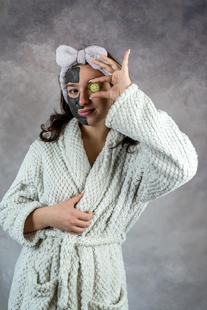 junge Frau in weißem Bademantel, die Gurkenschnitte hält, schwarze Lehmmaske auf das Gesicht aufträgt, isoliert auf grauem Gesichtspflegeverfahren