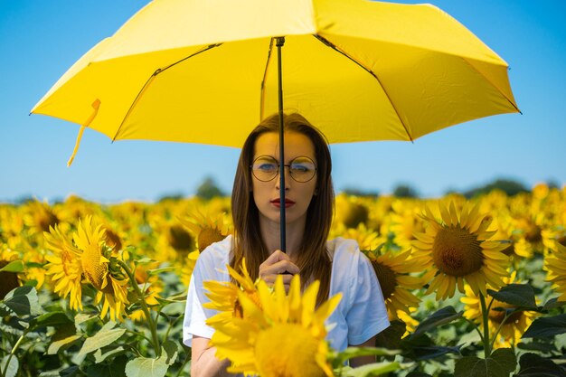 Junge Frau in weißem T-Shirt und Brille unter einem gelben Regenschirm auf einem Sonnenblumenfeld